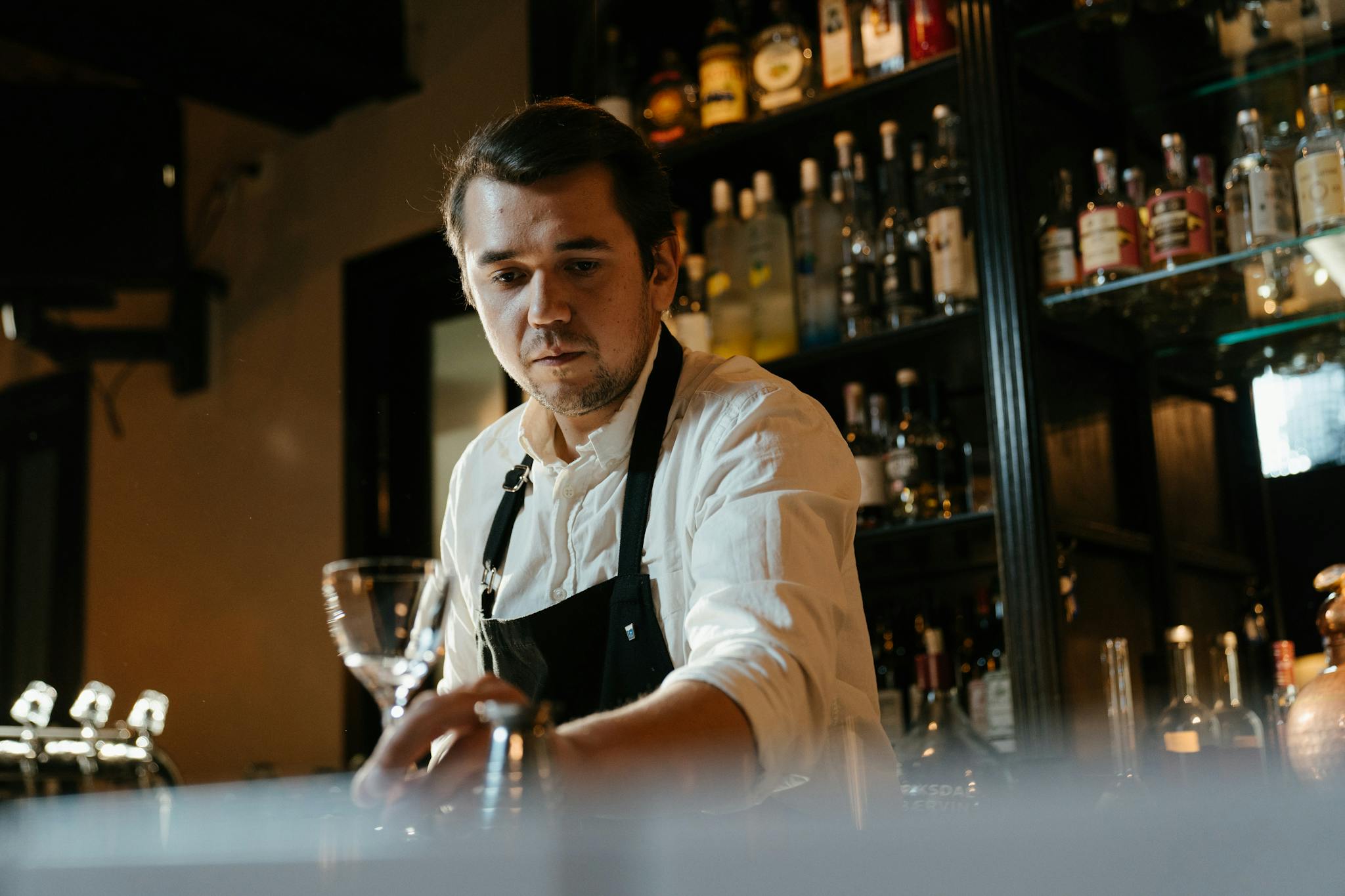 A skilled bartender in a white shirt and apron expertly crafting a cocktail in a dimly lit bar setting.